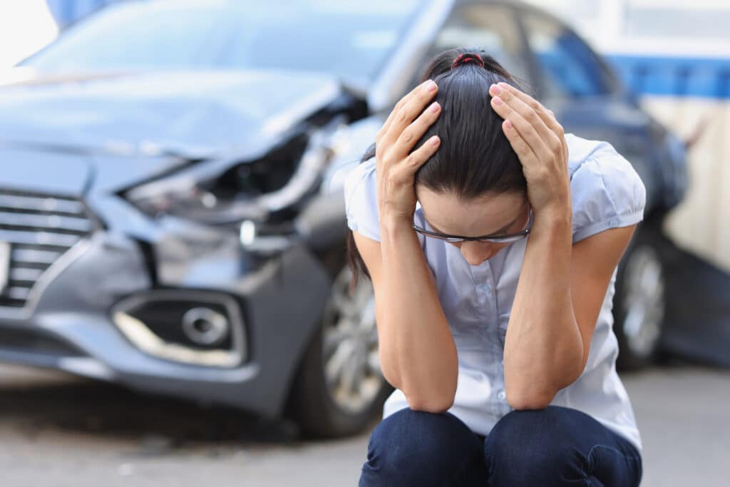 A Woman At A Broken Car Sits Holding Her Head, Close-up, Blurry.